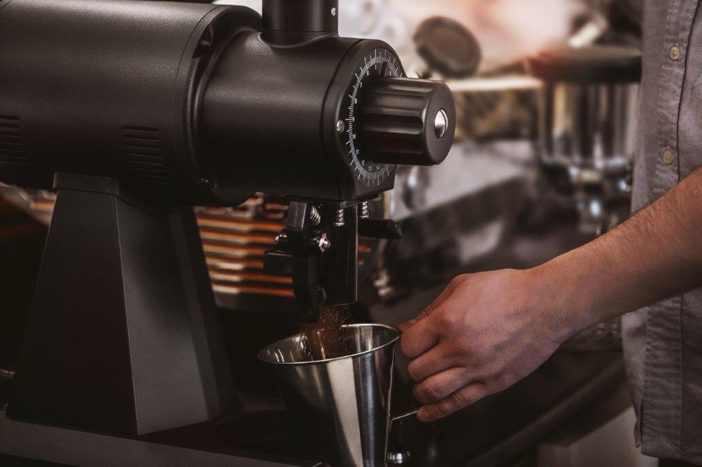 Barista mieli ziarna kawy do stalowego naczynia za pomocą profesjonalnego młynka Mahlkönig EK43. Widoczna jest charakterystyczna pokrętło regulacji grubości mielenia oraz przemysłowy design urządzenia, typowy dla kawiarni specialty i palarni. Zdjęcie oddaje precyzję i jakość mielenia oferowaną przez niemieckiego producenta młynków klasy premium.
