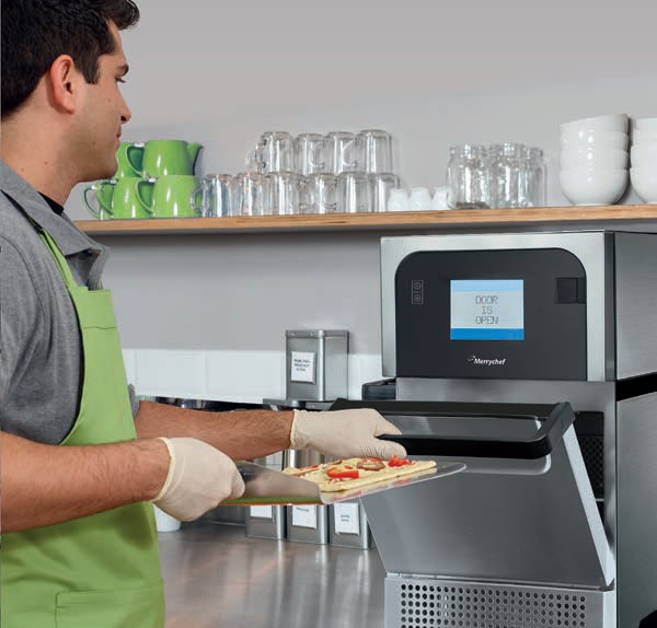 Pracownik gastronomii wkłada potrawę do pieca konwekcyjno-mikrofalowego Merrychef w nowoczesnym zapleczu kuchennym. Na zdjęciu widać profesjonalny piec szybkokomorowy, otwartą komorę oraz stanowisko pracy z naczyniami i akcesoriami barowymi w tle. Ujęcie prezentuje ergonomię, higienę i efektywność pracy z piecem Merrychef, idealnym do lokali typu kawiarnia, bistro i fast-casual.